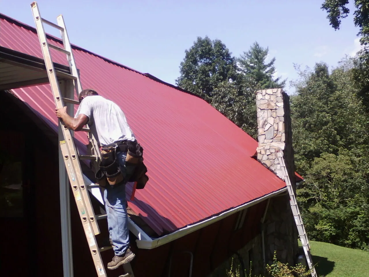 Roofer on a ladder beside a red metal roof and stone chimney — Soffit & Fascia Repair by Roofing Brothers in Lowry City